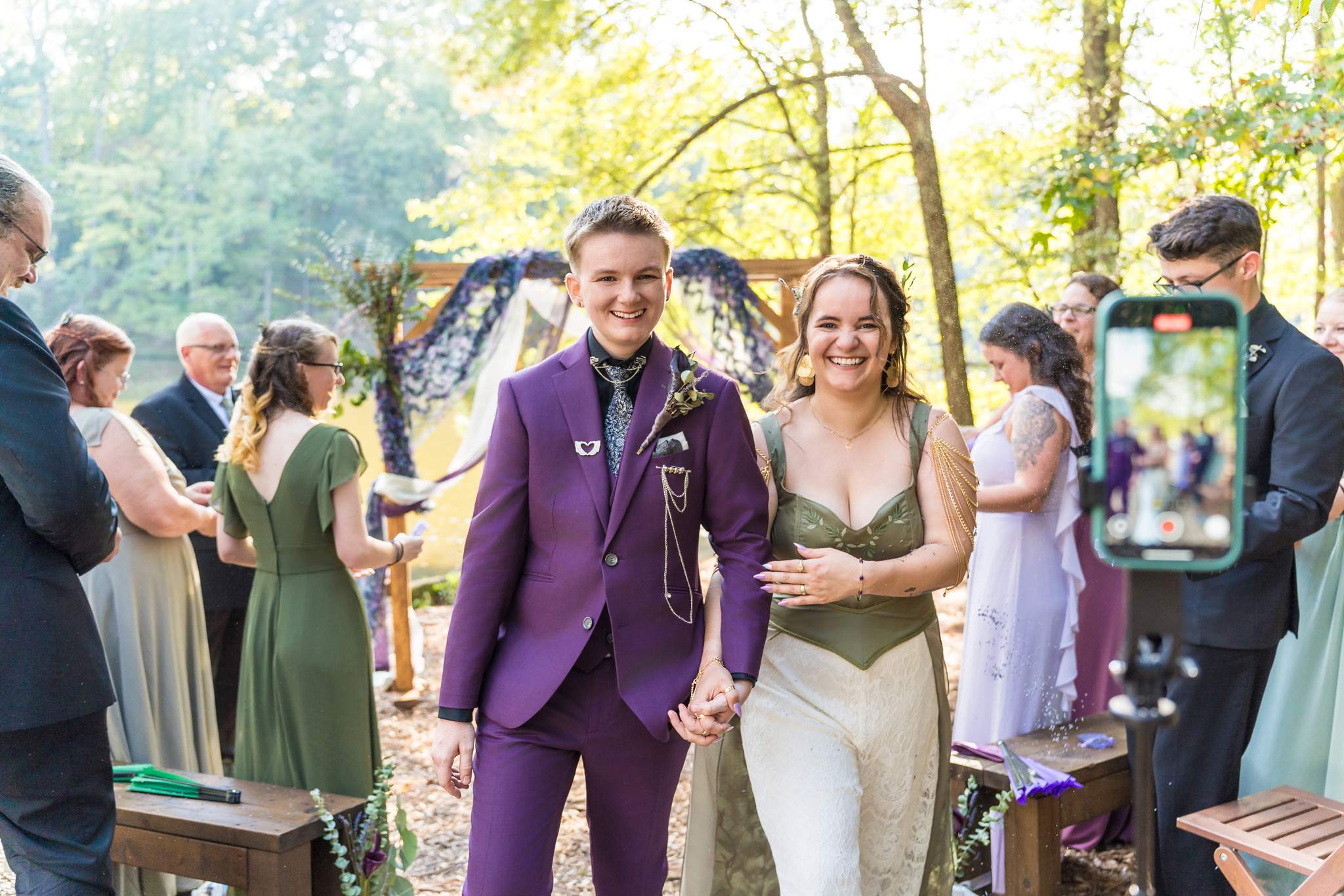 Purple suit and sage corset with cream pants as the couple walks out past guests in green