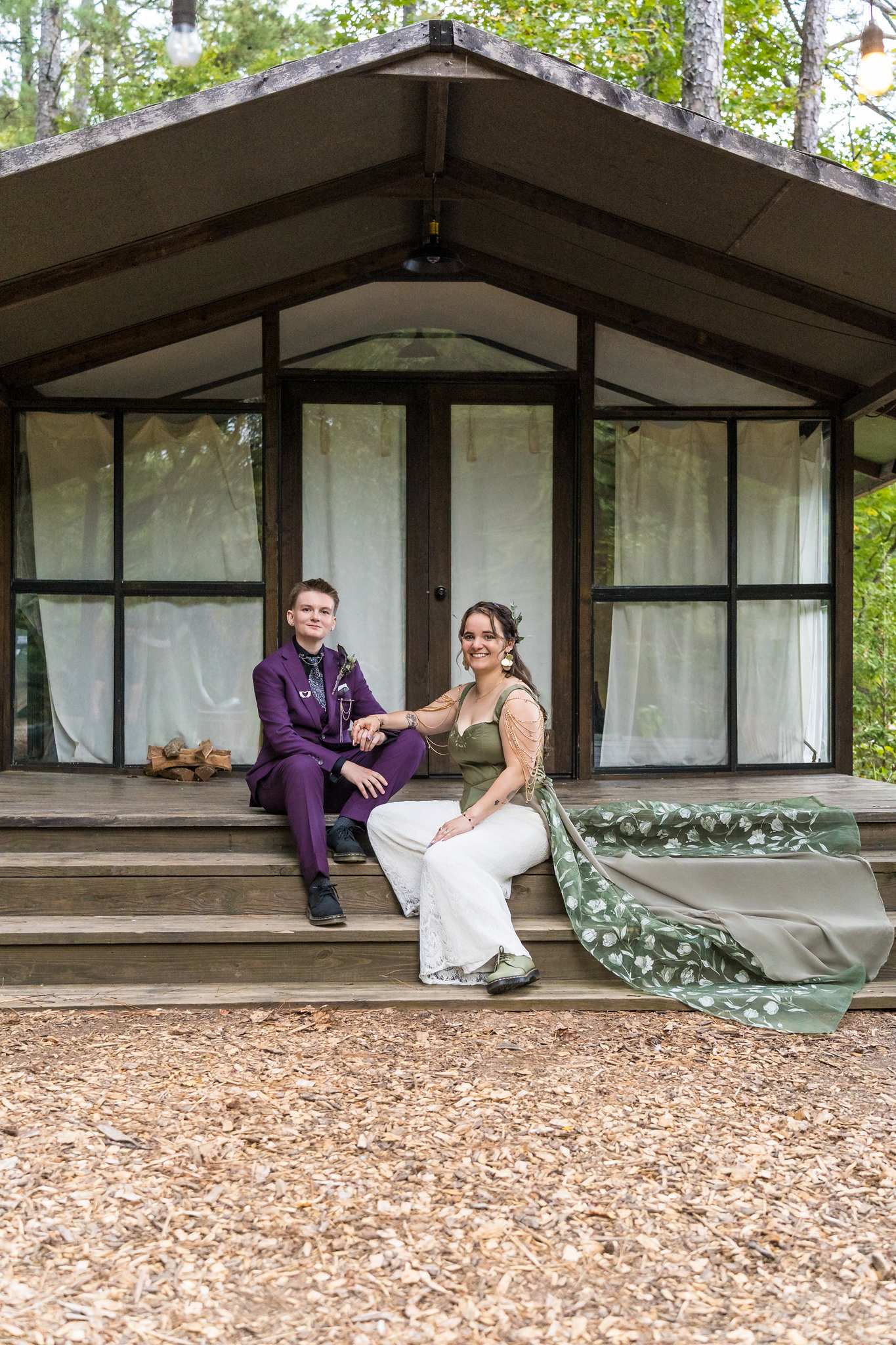 Couple in a purple suit and a sage corset with cream wide-leg pants sitting on rustic cabin steps