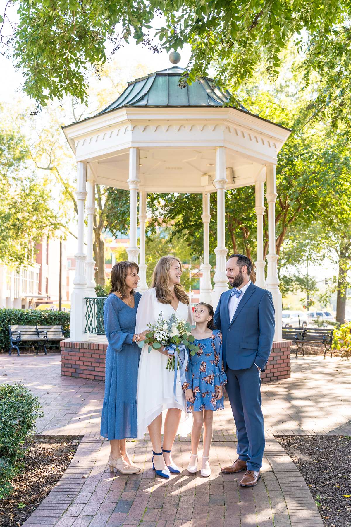 Bride in a white midi dress with navy heels, family in dusty blue, by a white gazebo