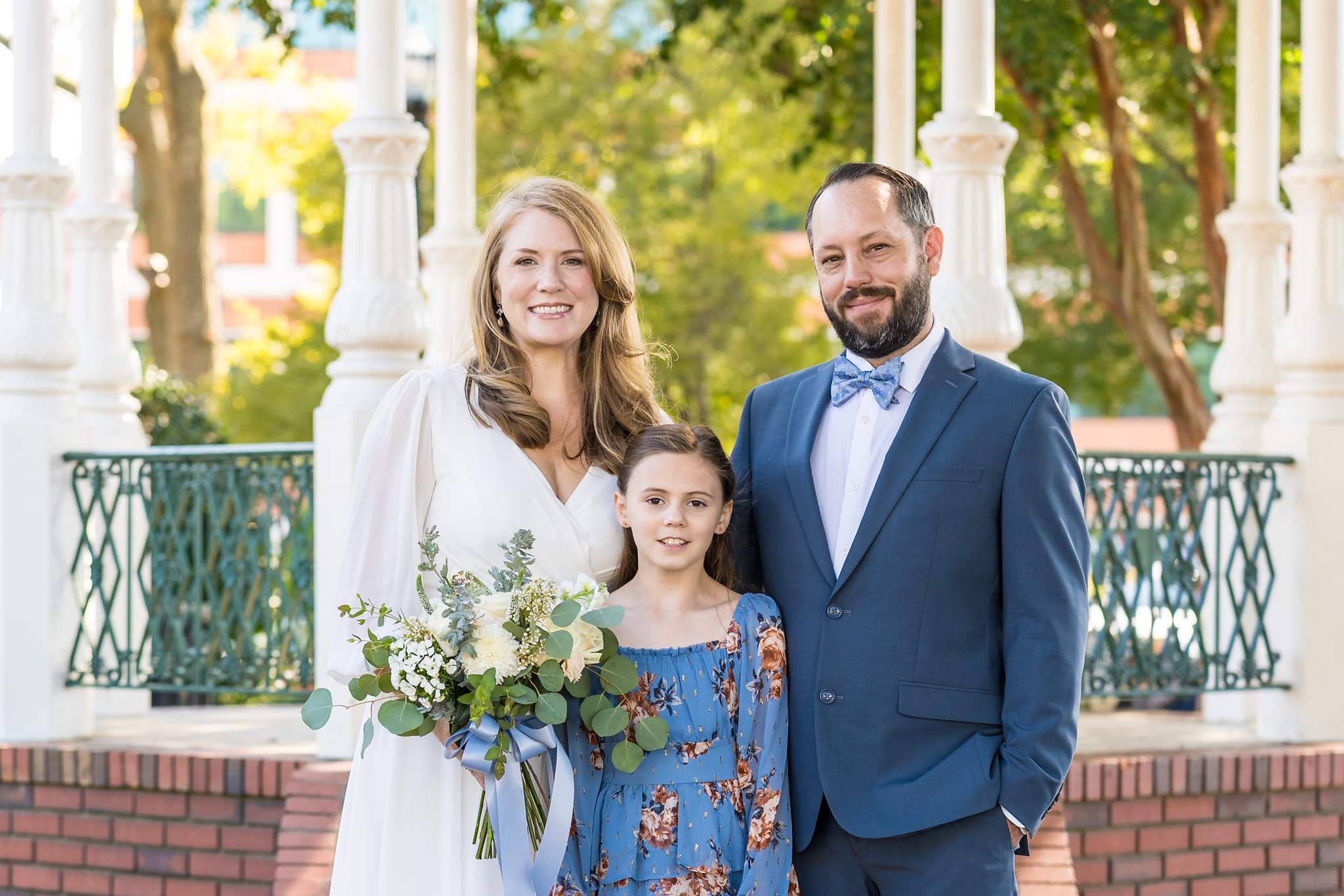 Couple in a blue suit and white chiffon dress with a girl in blue florals at a gazebo