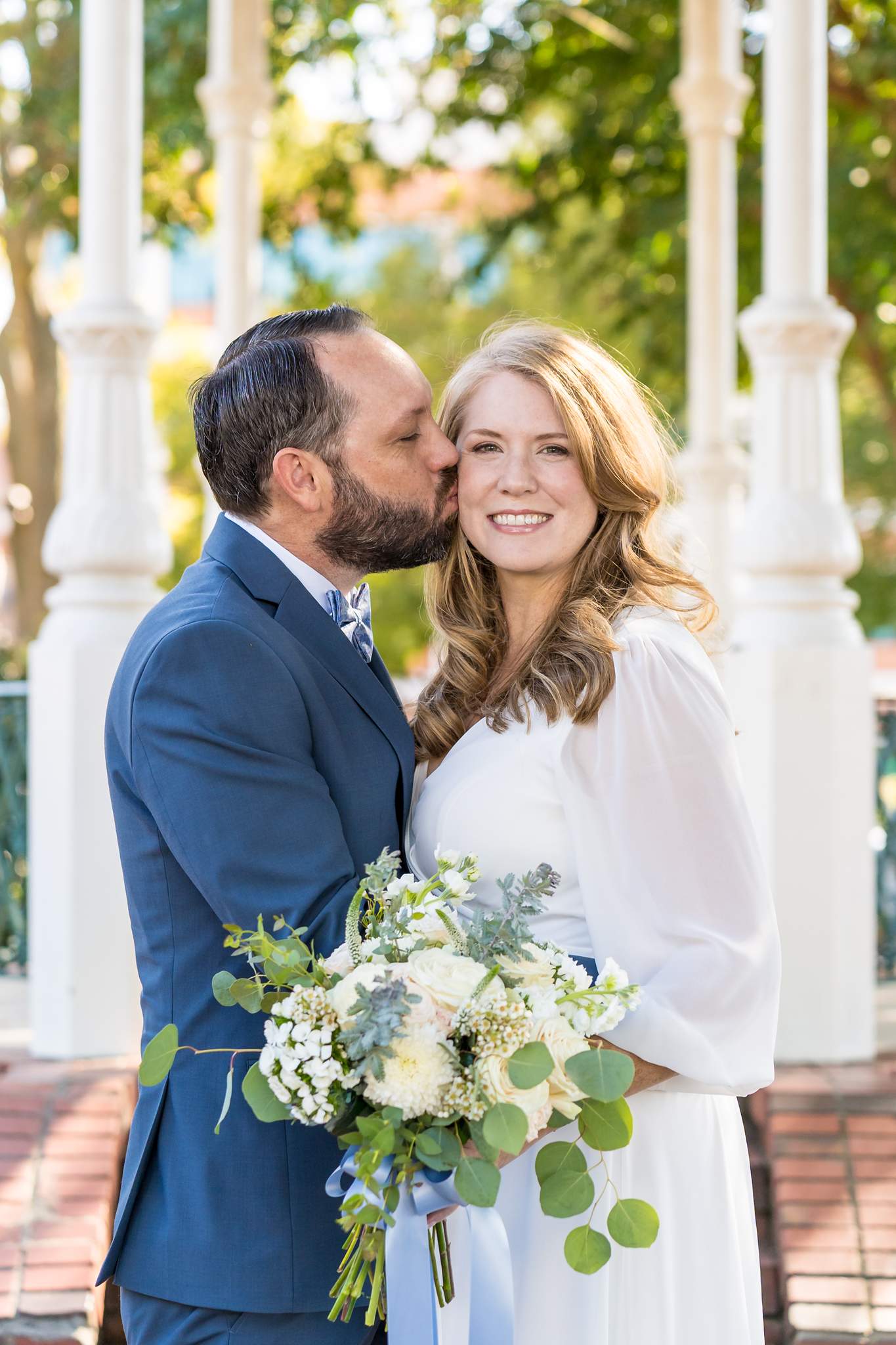 Groom in a blue suit and light blue bow tie kisses the bride in sheer white sleeves