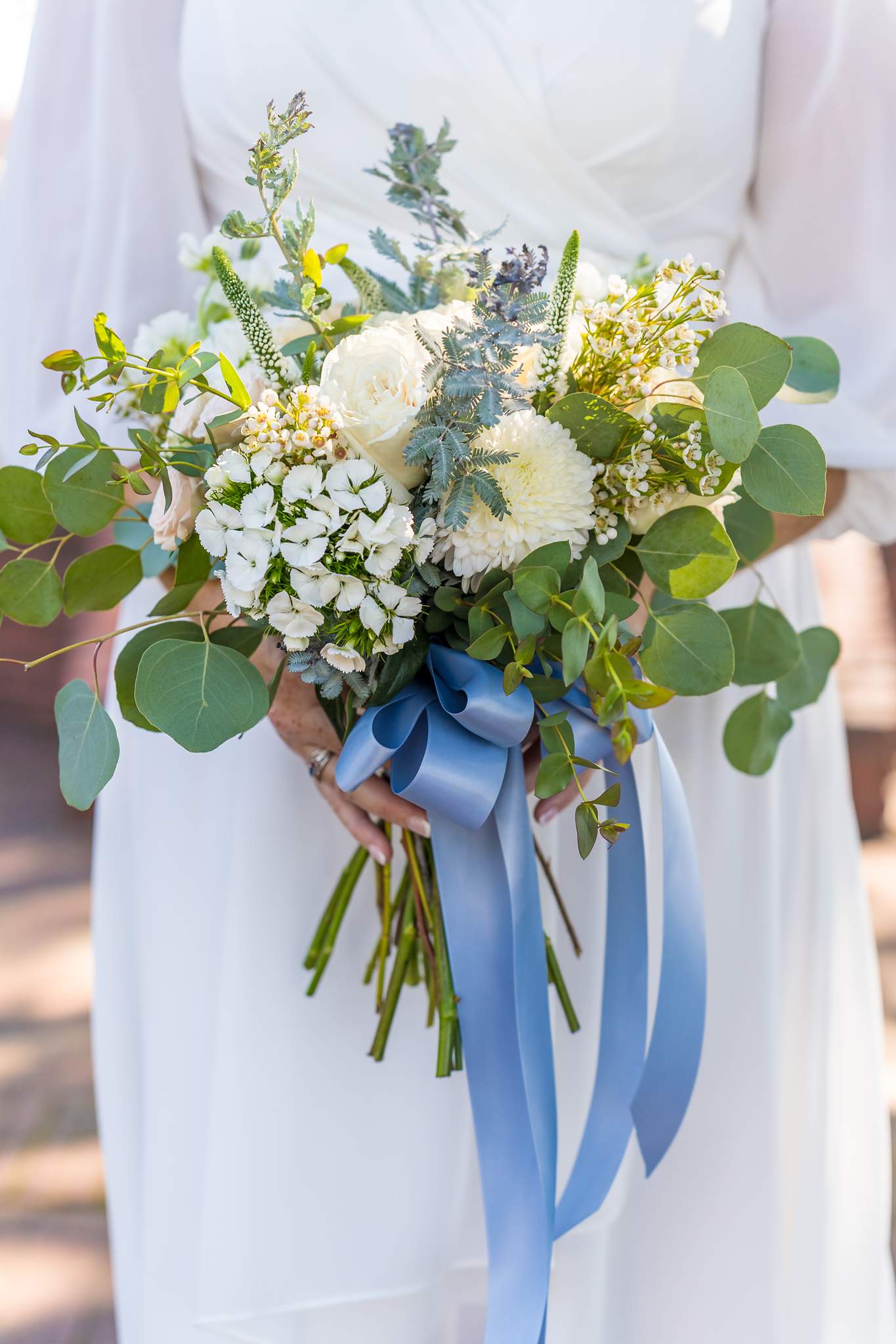 White flowers and eucalyptus bouquet tied with long dusty blue satin ribbons against a white chiffon dress