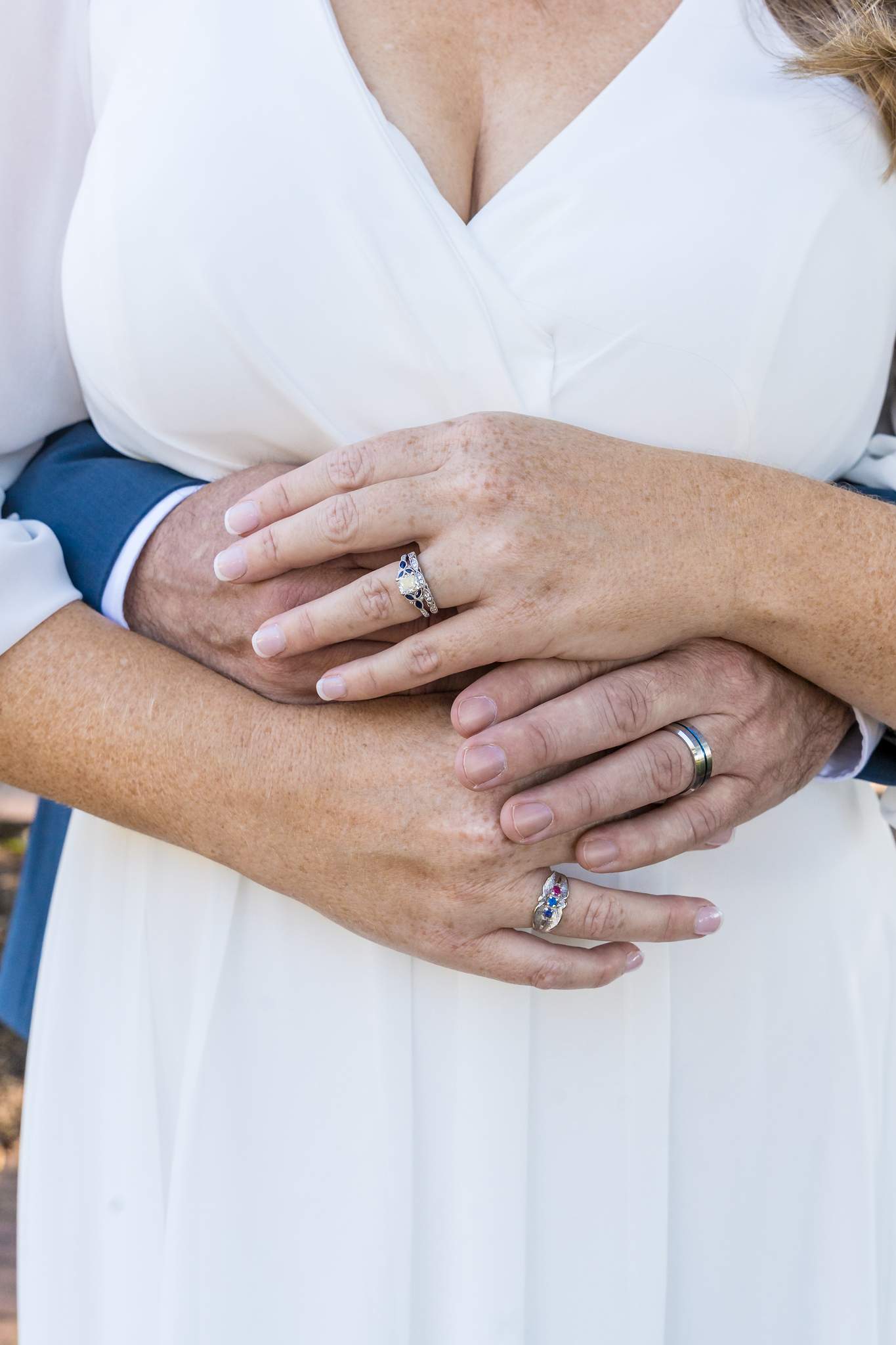 Wedding rings on clasped hands over the bride's white wrap-style chiffon dress and the groom's blue sleeve