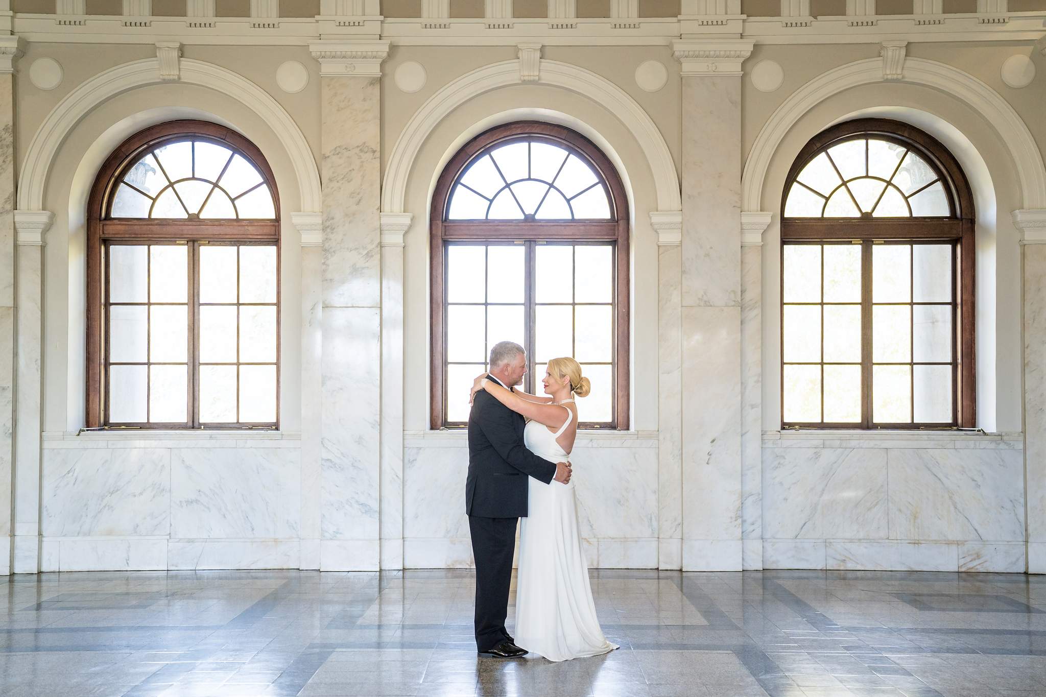 Couple in a white gown and black tuxedo embracing beneath tall arched windows in a marble courthouse hall