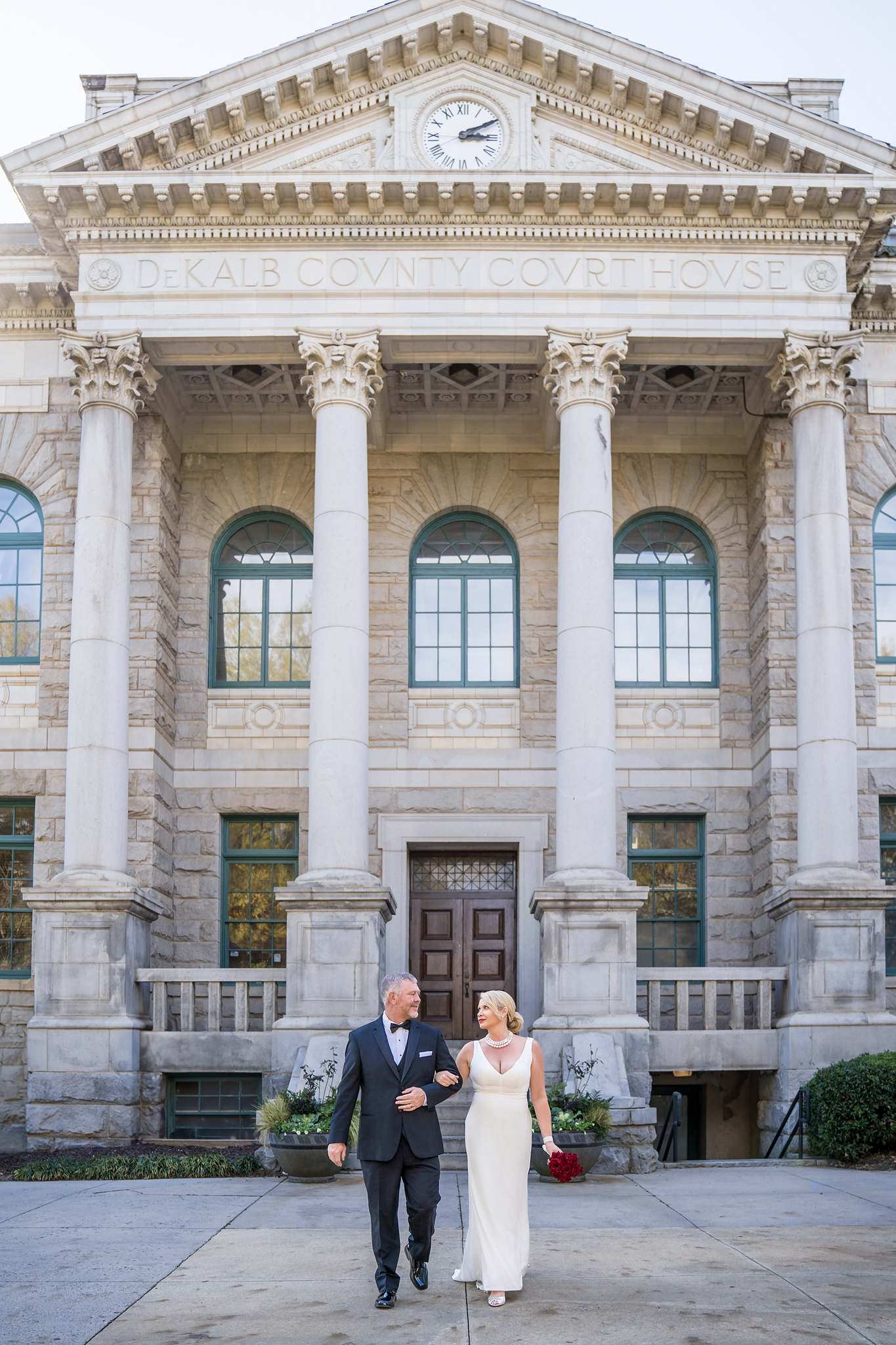 Groom in a black tuxedo and bride in a white gown walking out from a columned historic courthouse