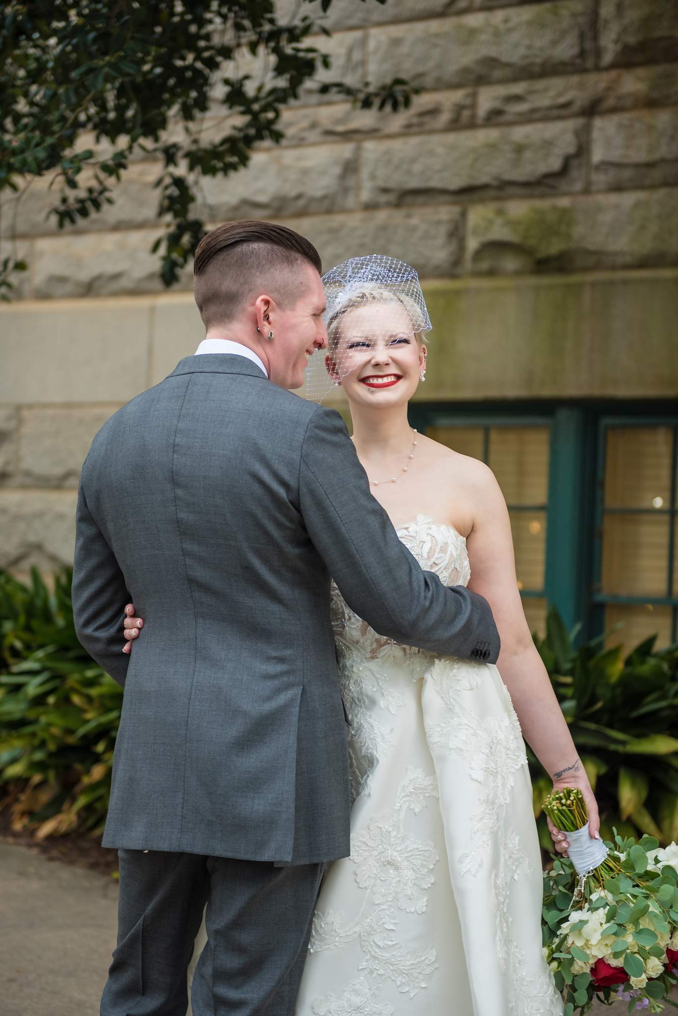 Groom in a gray suit hugging the bride in a strapless embroidered gown and short birdcage veil