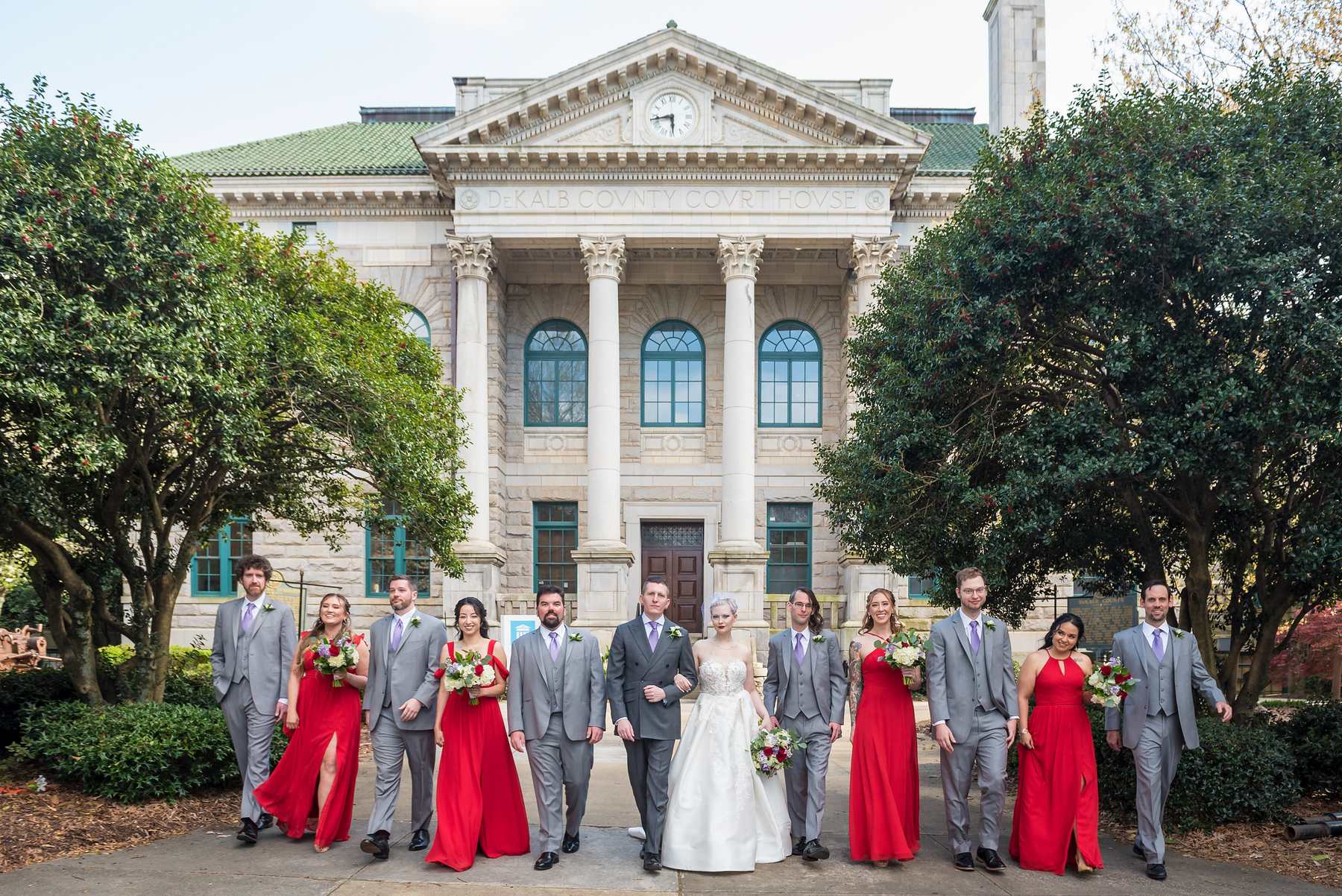 Bridesmaids in red chiffon gowns and groomsmen in gray suits walking with the couple outside a columned courthouse