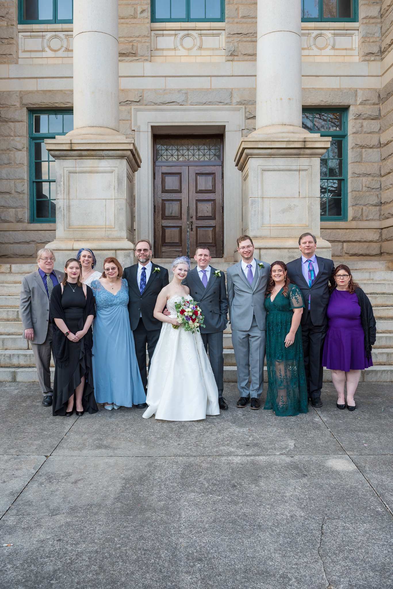 Family in black, dusty blue, emerald, purple and gray suits around the couple on stone courthouse steps