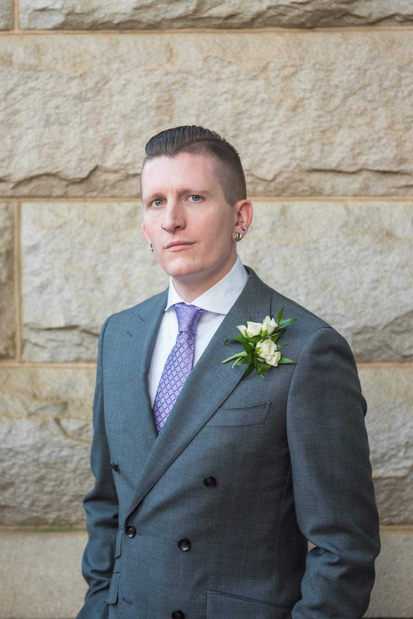 Groom in a gray double-breasted suit with a lavender tie and white flower boutonniere by stone walls