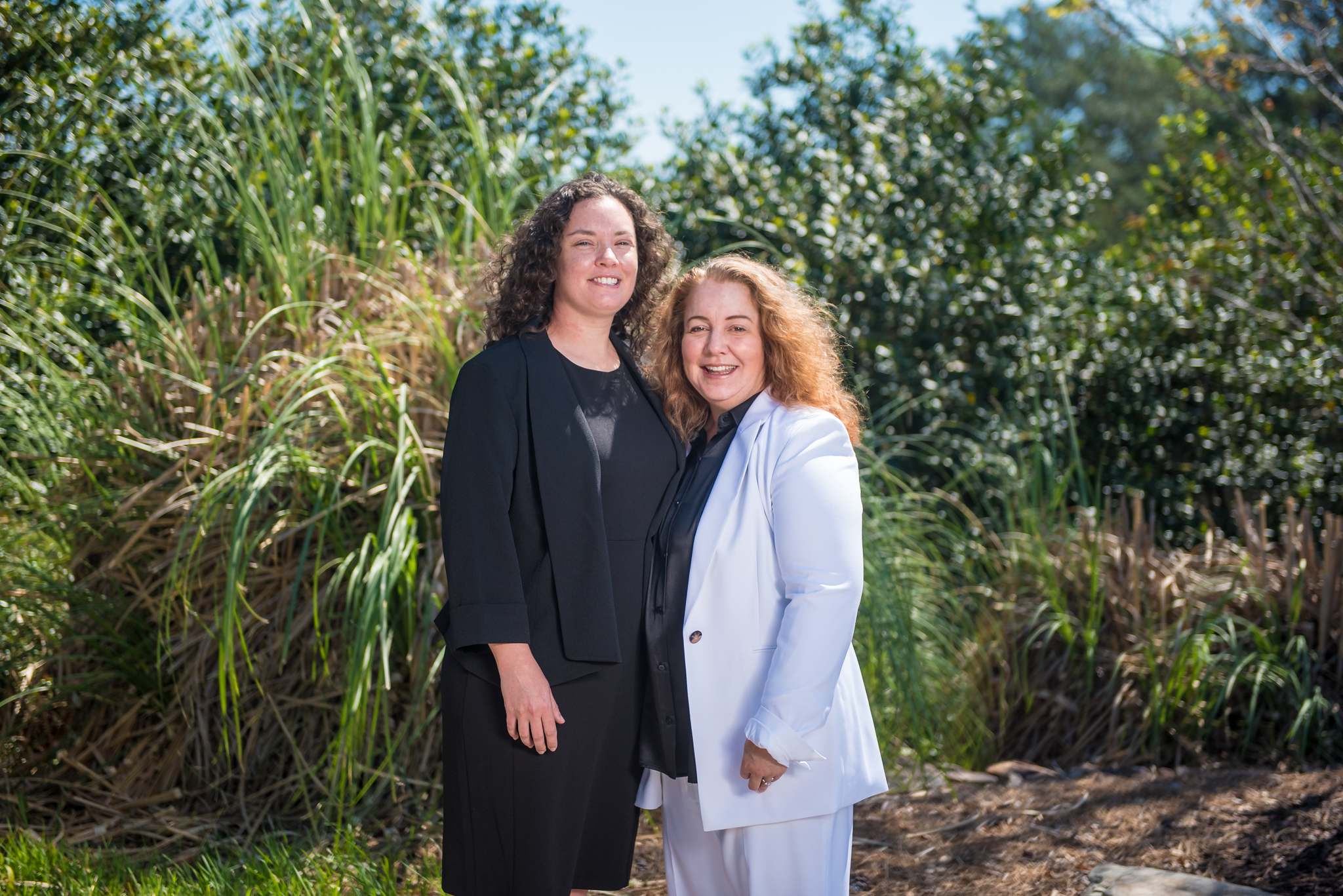 Couple in a black blazer and a white pantsuit on their wedding day among tall garden grasses