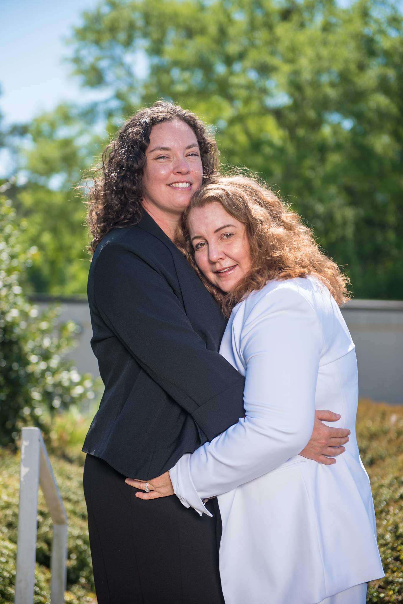 Couple in a black blazer and a white blazer suit hugging in a sunny garden