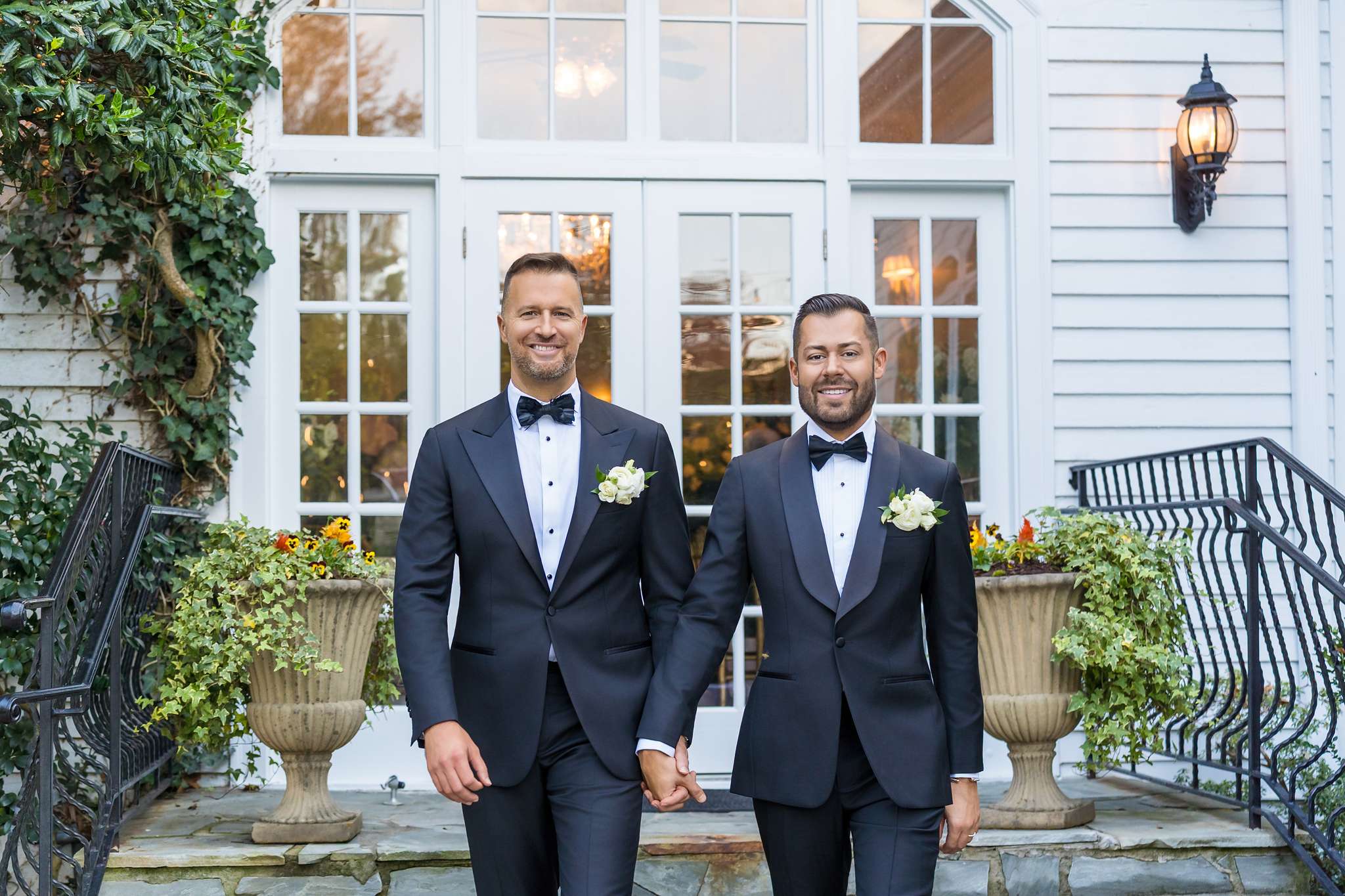 Two grooms holding hands in navy tuxedos with black bow ties and white rose boutonnieres by French doors