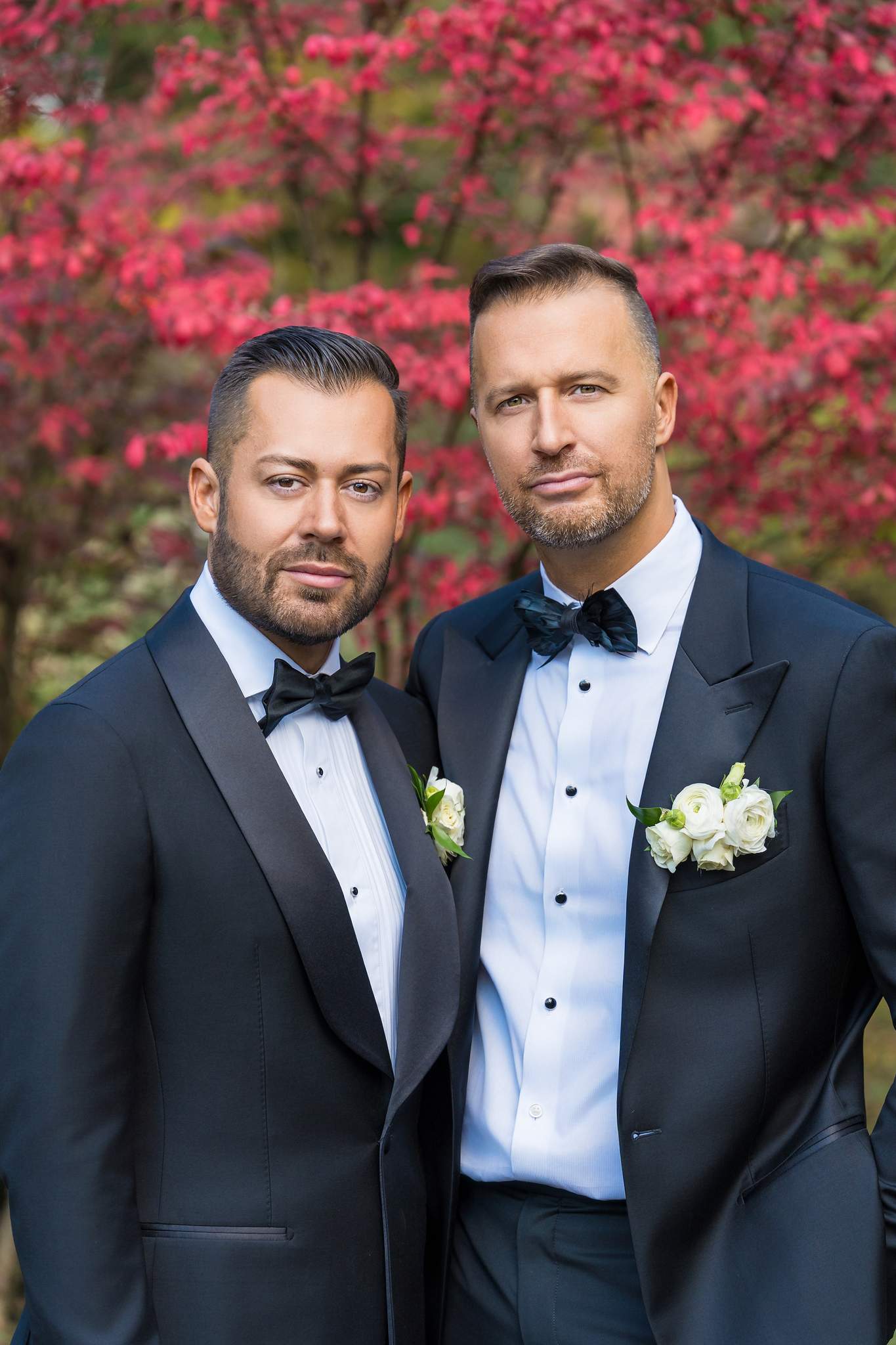 Two grooms in navy tuxedos with black bow ties and white rose boutonnieres before bright red fall leaves